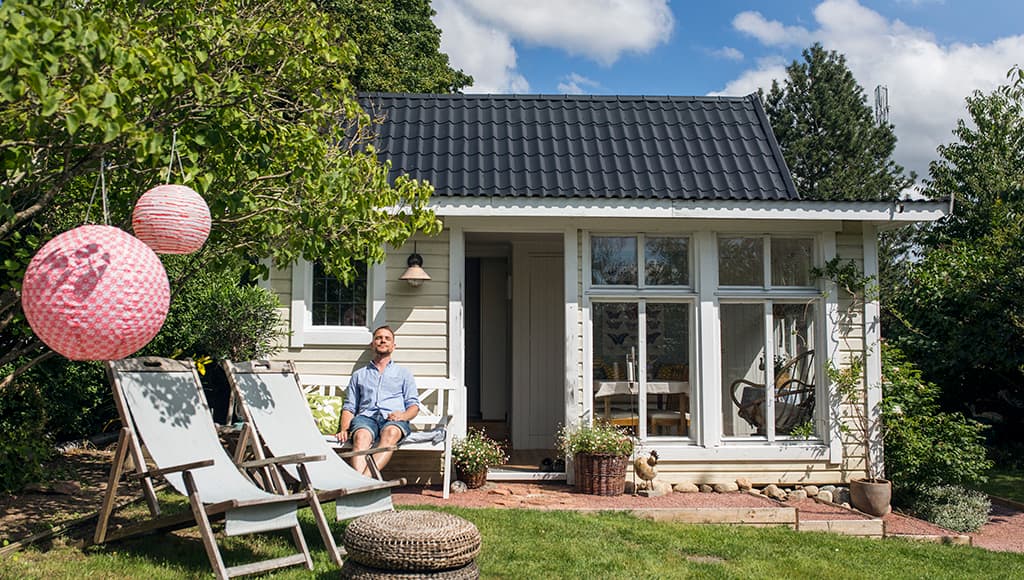 A man relaxes in front of a small cottage with large, sunny windows on a nice day