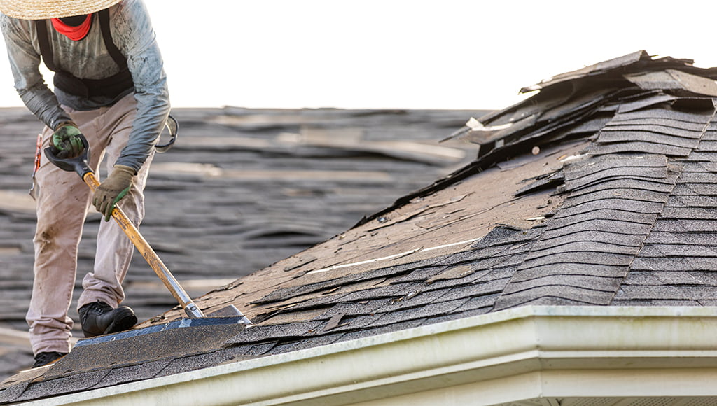 A roofer removes shingles during a roof repair. Edge flashing and gutters are visible.