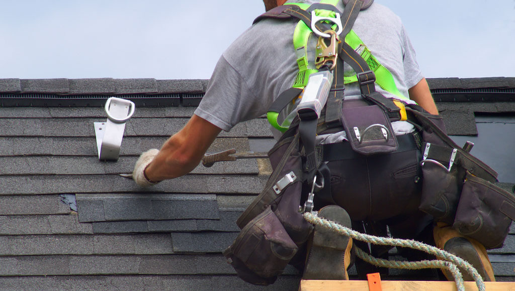 A roofer sits on a wooden brace while hammering nails into a shingle during a repair.